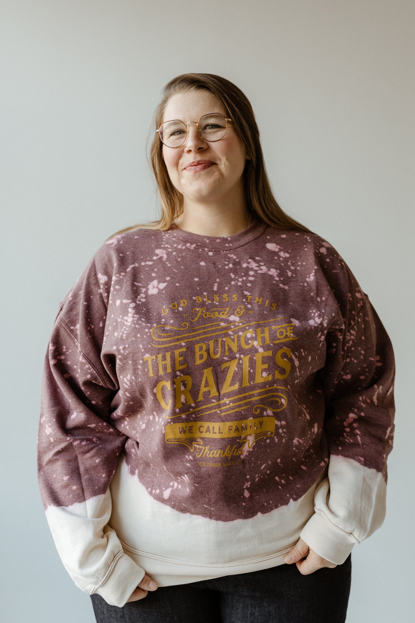 A person wearing glasses and a hand-bleached sweatshirt with a text design stands against a plain background, radiating Thanksgiving spirit.