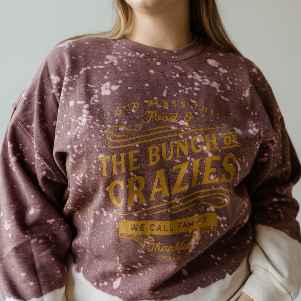 A woman wearing glasses and a purple, hand-bleached sweatshirt with gold text that reads, "Food & this bunch of crazies we call family, thankful," exudes the thanksgiving spirit as she stands against a plain background.