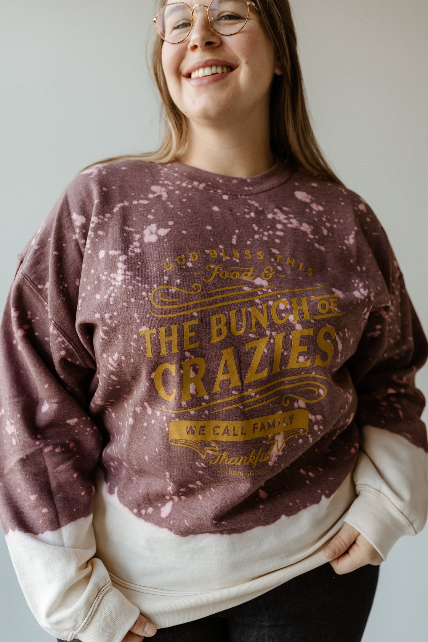 A woman wearing glasses and a purple, hand-bleached sweatshirt with gold text that reads, "Food & this bunch of crazies we call family, thankful," exudes the thanksgiving spirit as she stands against a plain background.