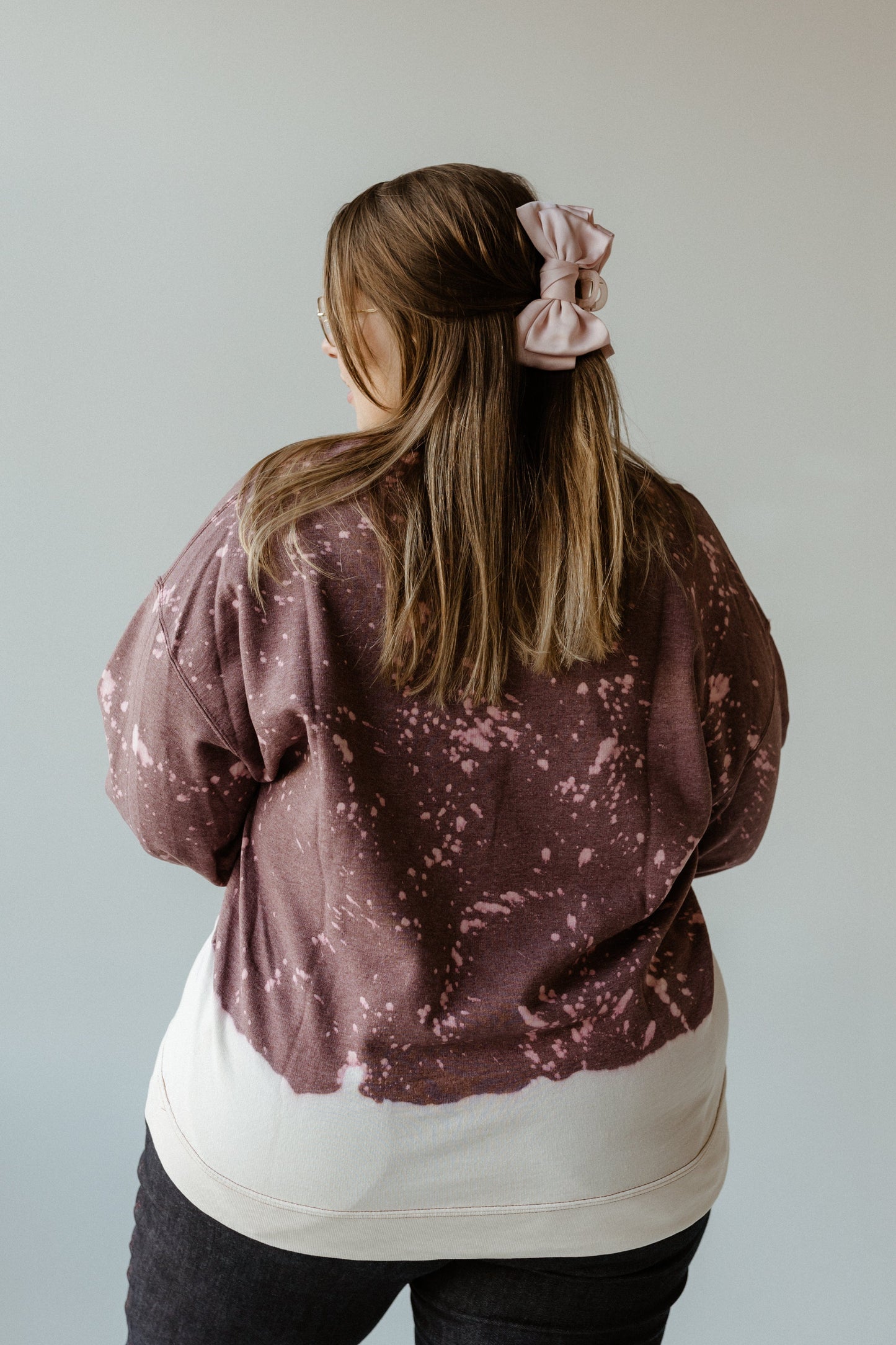 A person with long hair is seen from the back, donning a hand-bleached brown and pink tie-dye sweatshirt paired with a pink hair bow, exuding a festive Thanksgiving spirit.