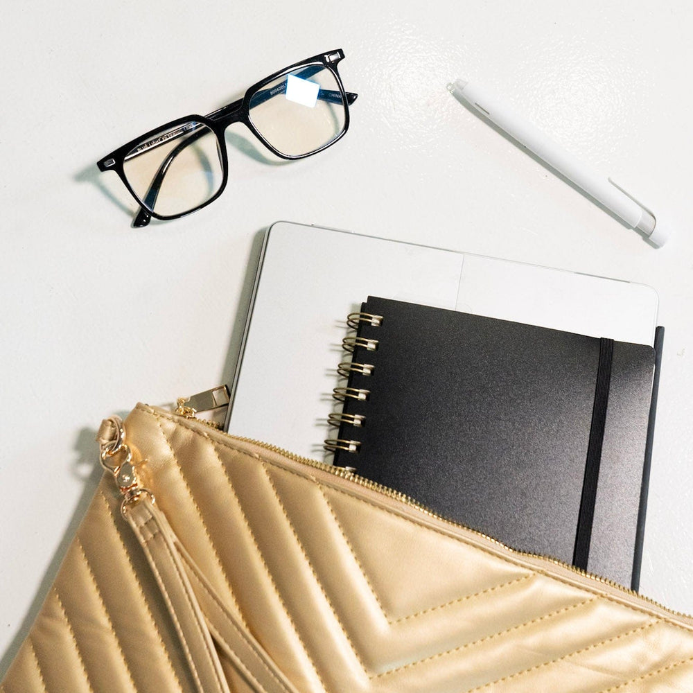 Gold clutch, glasses, notebook, and pen on a white surface