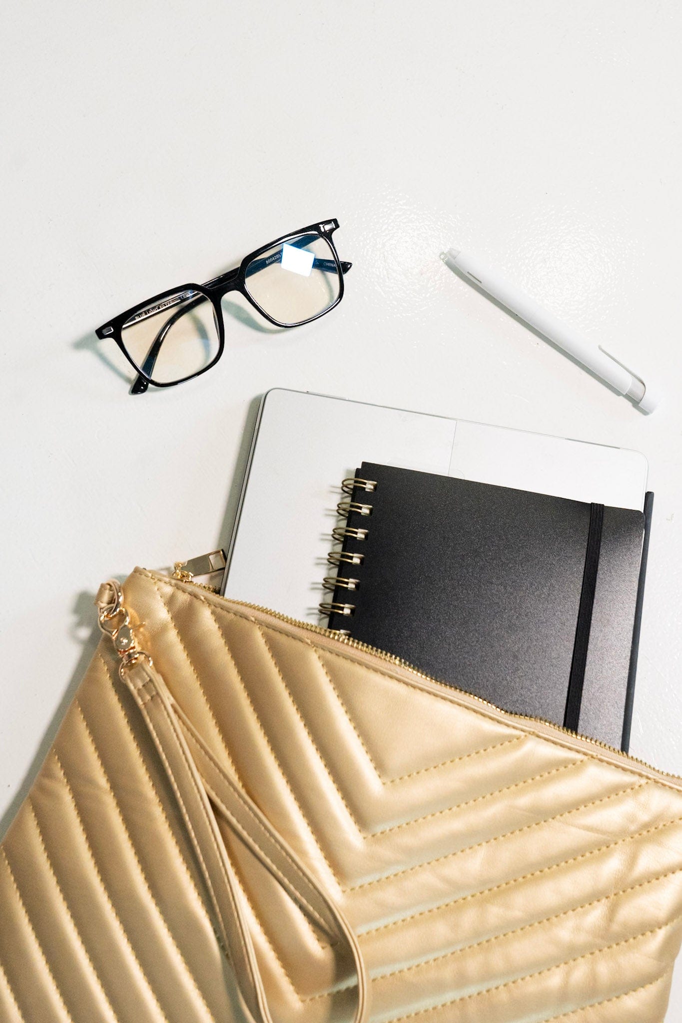 Gold clutch, glasses, notebook, and pen on a white surface