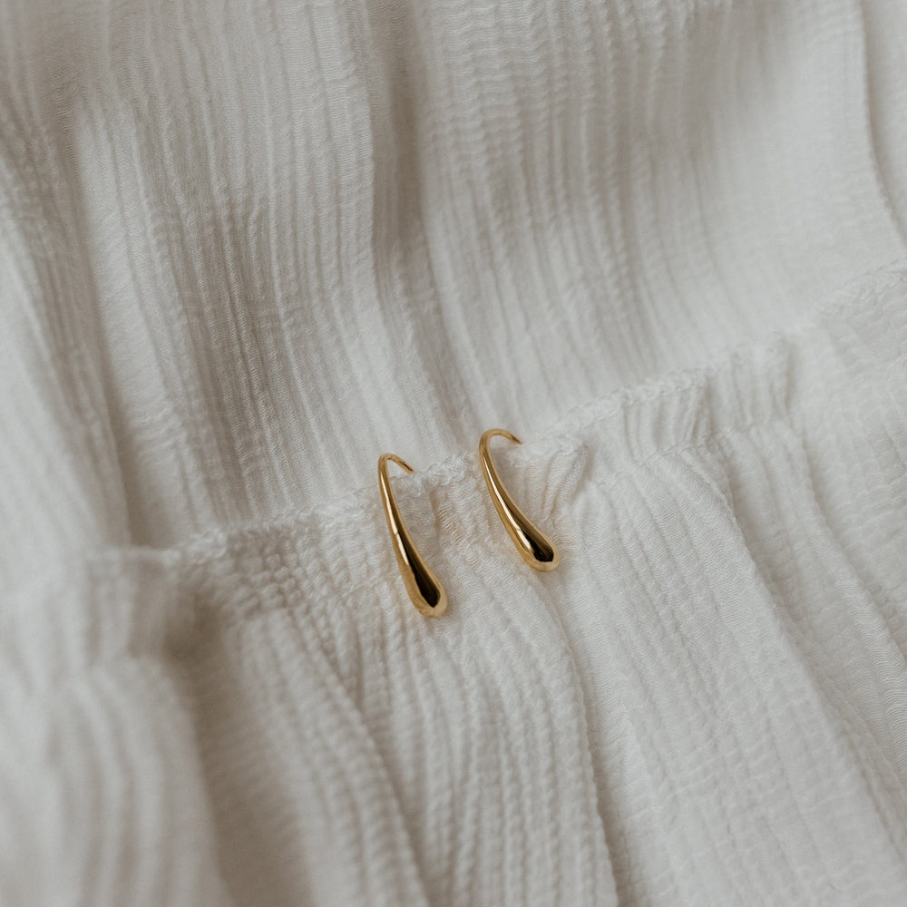 The GRACE EARRINGS, meticulously crafted in 18k gold, gently curve as they rest on a textured white fabric surface.