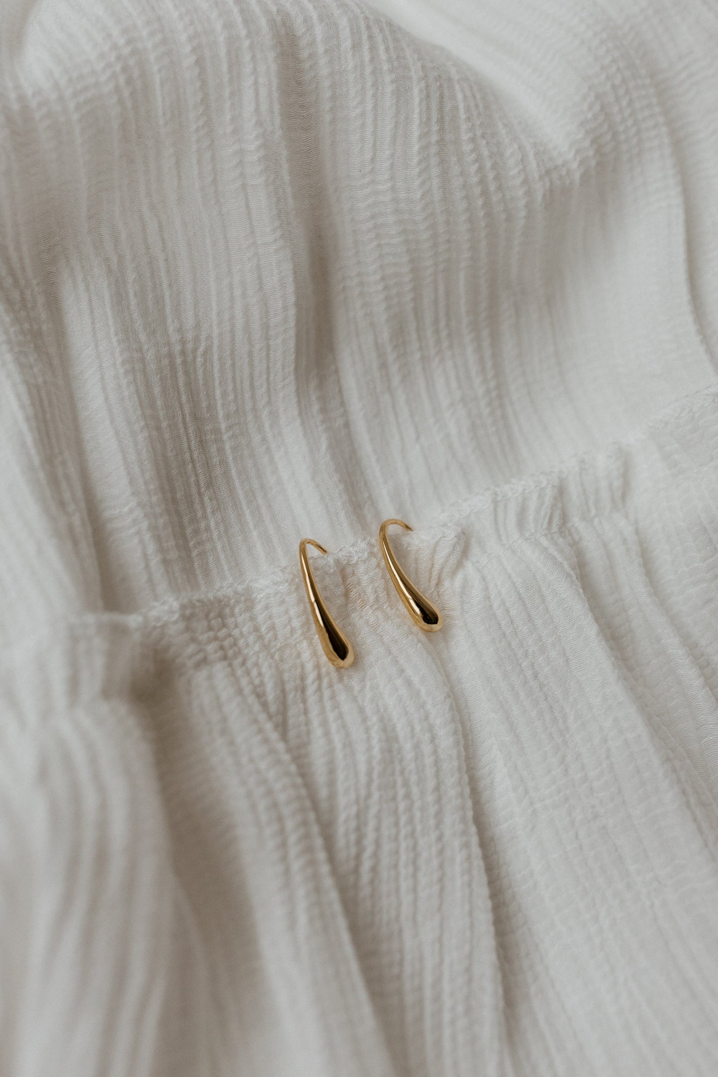 The GRACE EARRINGS, meticulously crafted in 18k gold, gently curve as they rest on a textured white fabric surface.