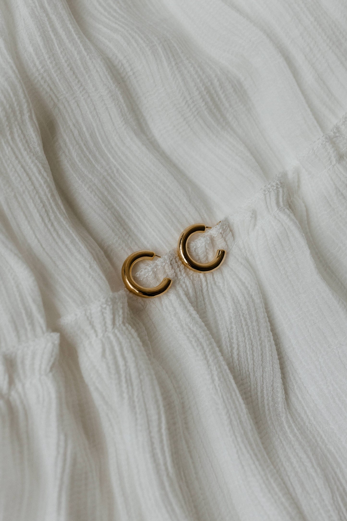 Close-up of a white textured fabric adorned with two small gold hoops from the GRETA HOOP EARRINGS collection.