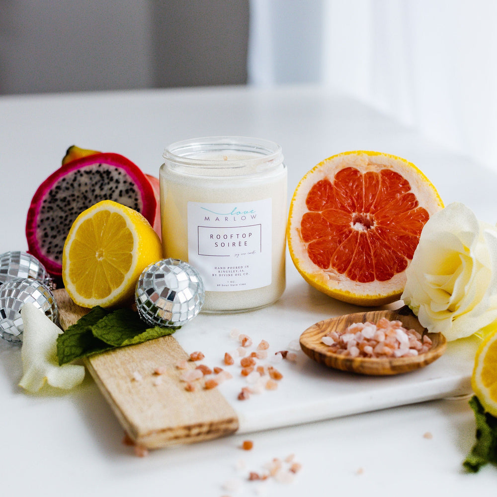 A jar candle labeled "HANDCRAFTED SOY WAX CANDLES IN EXCLUSIVE SCENT ROOFTOP SOIRÉE" by Love Marlow is placed on a white surface surrounded by a sliced lemon, grapefruit, dragon fruit, rose, wooden board, and small bowl of pink salt. Made from soy wax and infused with essential oils, it creates the perfect ambiance for a Rooftop Soirée.