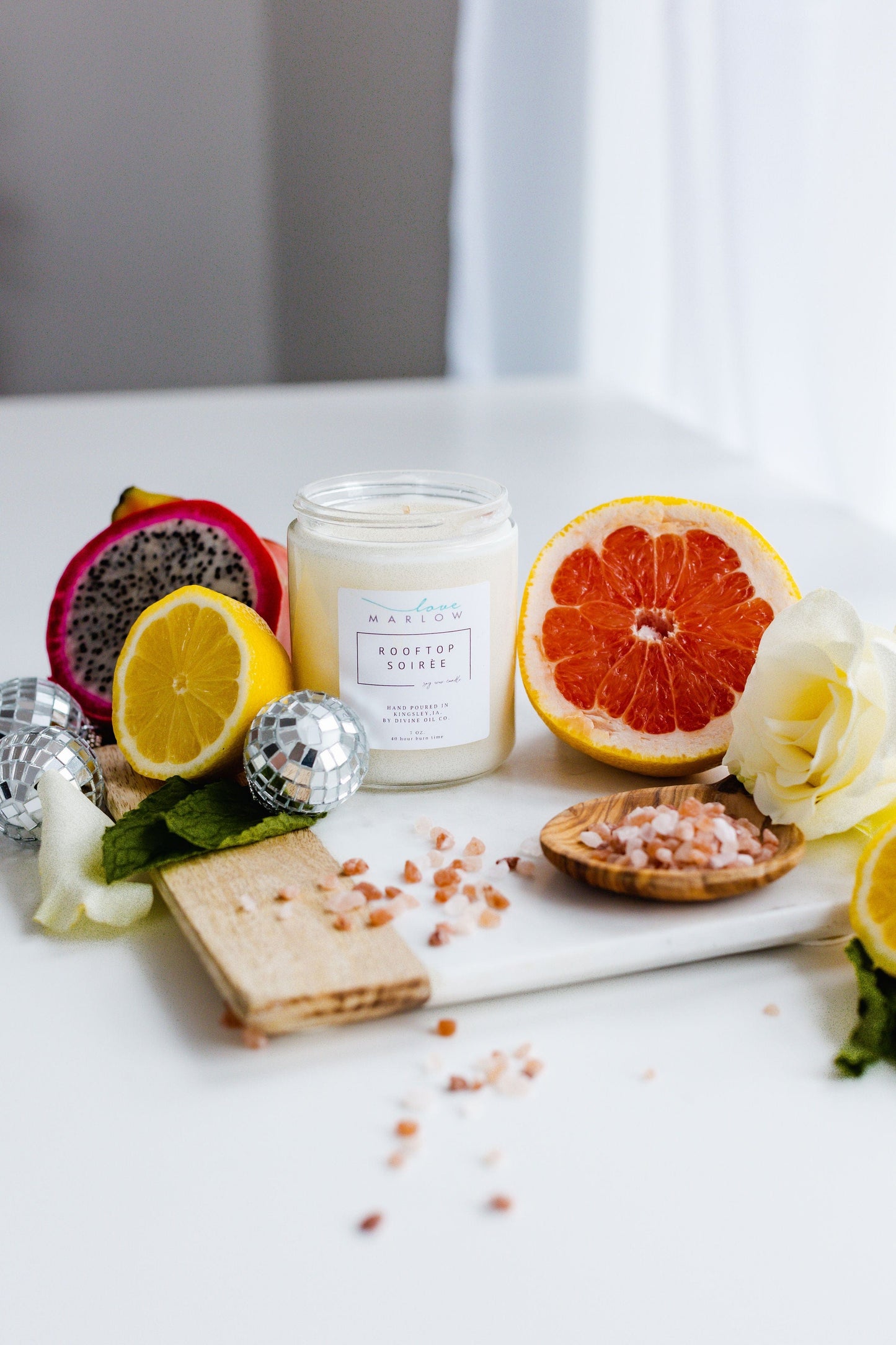 A jar candle labeled "HANDCRAFTED SOY WAX CANDLES IN EXCLUSIVE SCENT ROOFTOP SOIRÉE" by Love Marlow is placed on a white surface surrounded by a sliced lemon, grapefruit, dragon fruit, rose, wooden board, and small bowl of pink salt. Made from soy wax and infused with essential oils, it creates the perfect ambiance for a Rooftop Soirée.