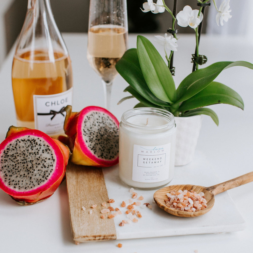 A table with a bottle of champagne, a filled champagne flute, sliced dragon fruit, a lit HANDCRAFTED SOY WAX CANDLES IN EXCLUSIVE SCENT WEEKEND GETAWAY by Love Marlow, a potted orchid, and a wooden spoon with pink salt.