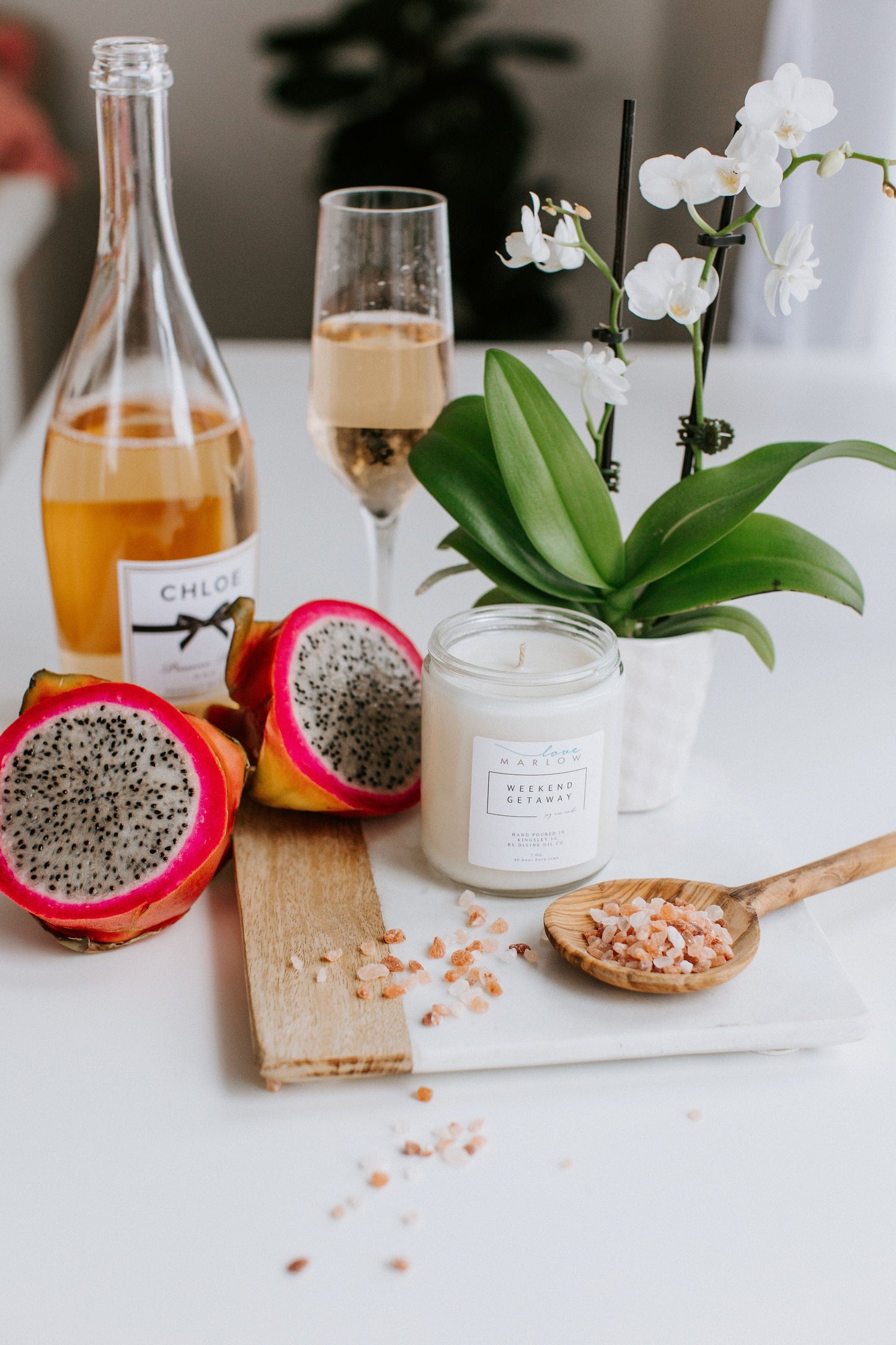 A table with a bottle of champagne, a filled champagne flute, sliced dragon fruit, a lit HANDCRAFTED SOY WAX CANDLES IN EXCLUSIVE SCENT WEEKEND GETAWAY by Love Marlow, a potted orchid, and a wooden spoon with pink salt.