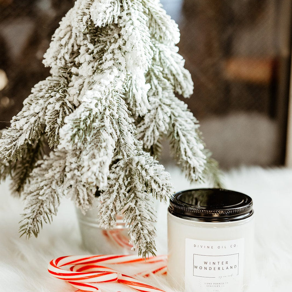 A handcrafted snow-covered miniature tree stands next to a lit soy wax candle labeled "Winter Wonderland," accompanied by two candy canes on a pristine white surface.