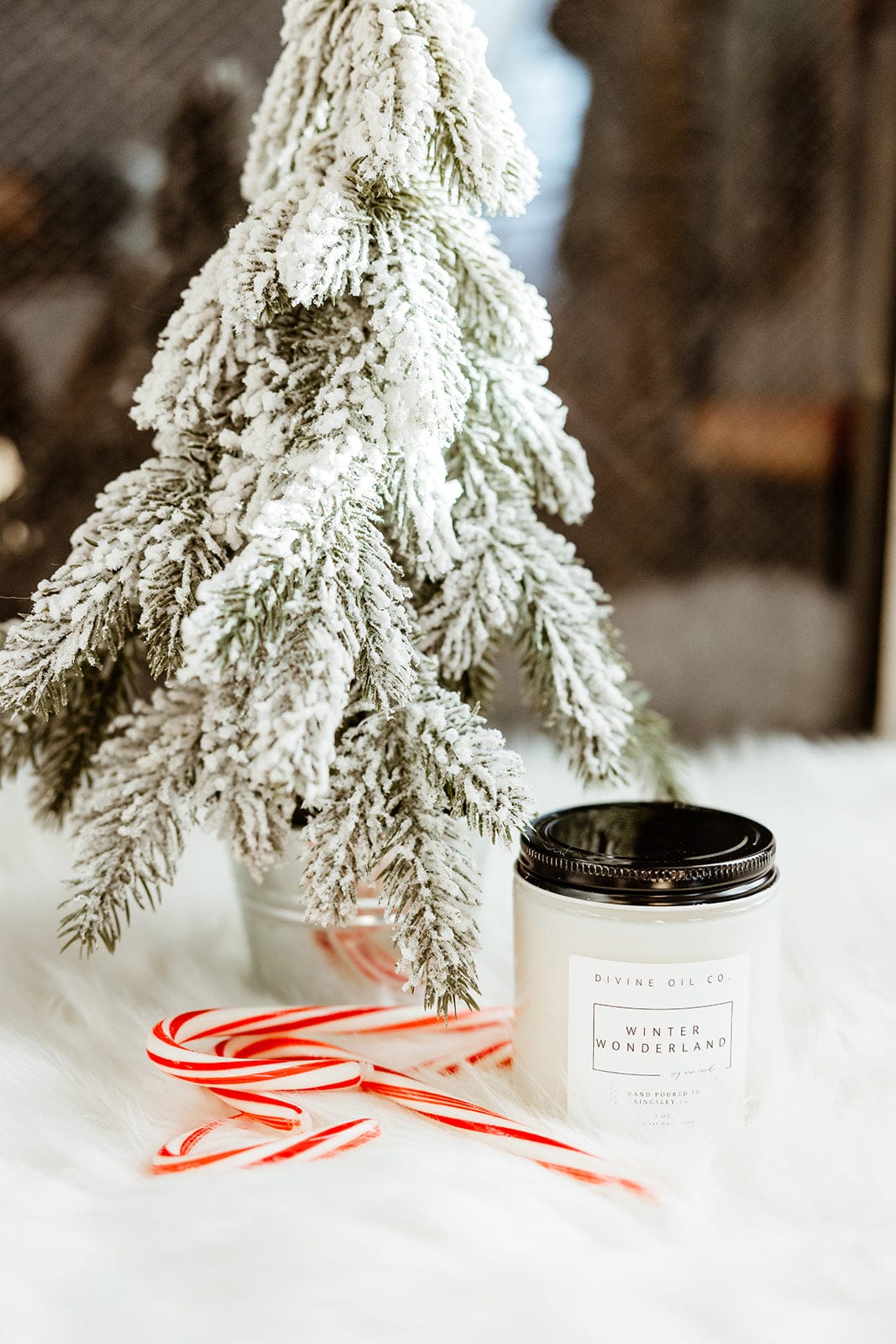 A handcrafted snow-covered miniature tree stands next to a lit soy wax candle labeled "Winter Wonderland," accompanied by two candy canes on a pristine white surface.