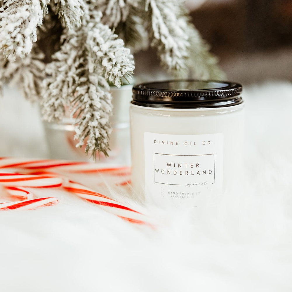 A handcrafted "Winter Wonderland" soy wax candle sits elegantly beside two candy canes and a snowy pine branch, all resting on a white, fluffy surface infused with the soothing aroma of essential oils.