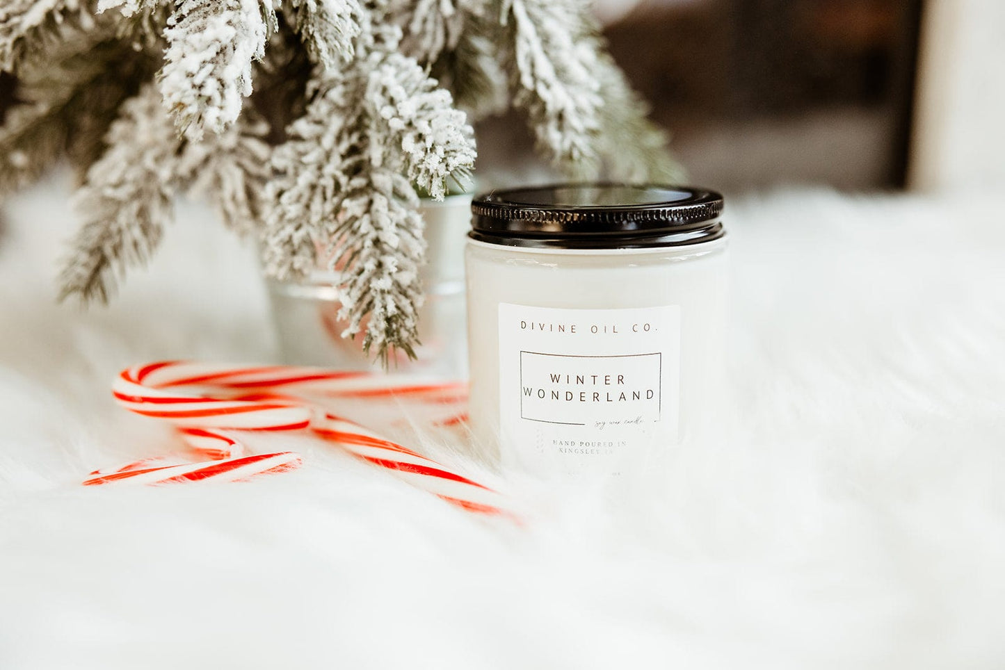 A handcrafted "Winter Wonderland" soy wax candle sits elegantly beside two candy canes and a snowy pine branch, all resting on a white, fluffy surface infused with the soothing aroma of essential oils.