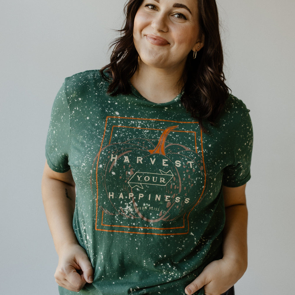 Wearing a Love Marlow "HARVEST YOUR HAPPINESS" GRAPHIC TEE in hand-bleached green, a person stands against a plain background, effortlessly highlighting fall fashion.