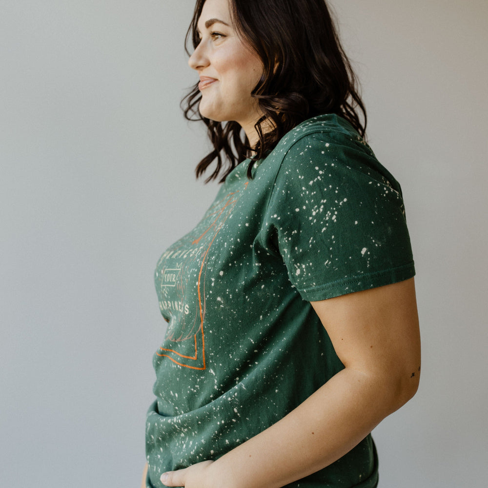 A person with shoulder-length dark hair in a "HARVEST YOUR HAPPINESS" GRAPHIC TEE by Love Marlow stands in profile, hands in pockets, against a plain background.