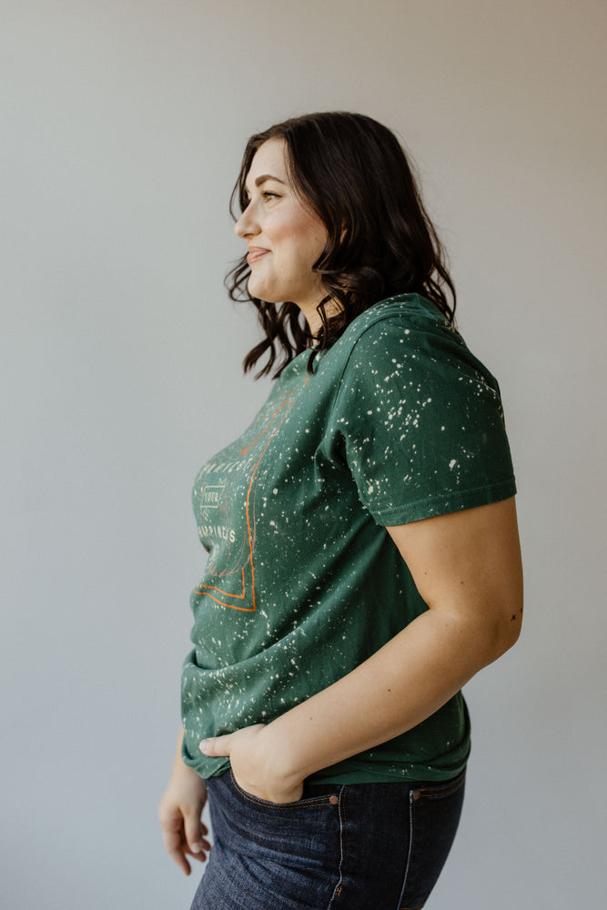 A person with shoulder-length dark hair in a "HARVEST YOUR HAPPINESS" GRAPHIC TEE by Love Marlow stands in profile, hands in pockets, against a plain background.