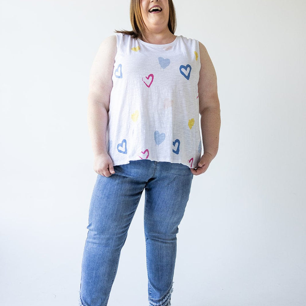 A person dressed in a sleeveless shirt adorned with colorful heart designs is standing and smiling against a plain background, wearing Love Marlow’s High-Rise Relaxed Skinny Jeans with Frayed Hem in Medium Wash.