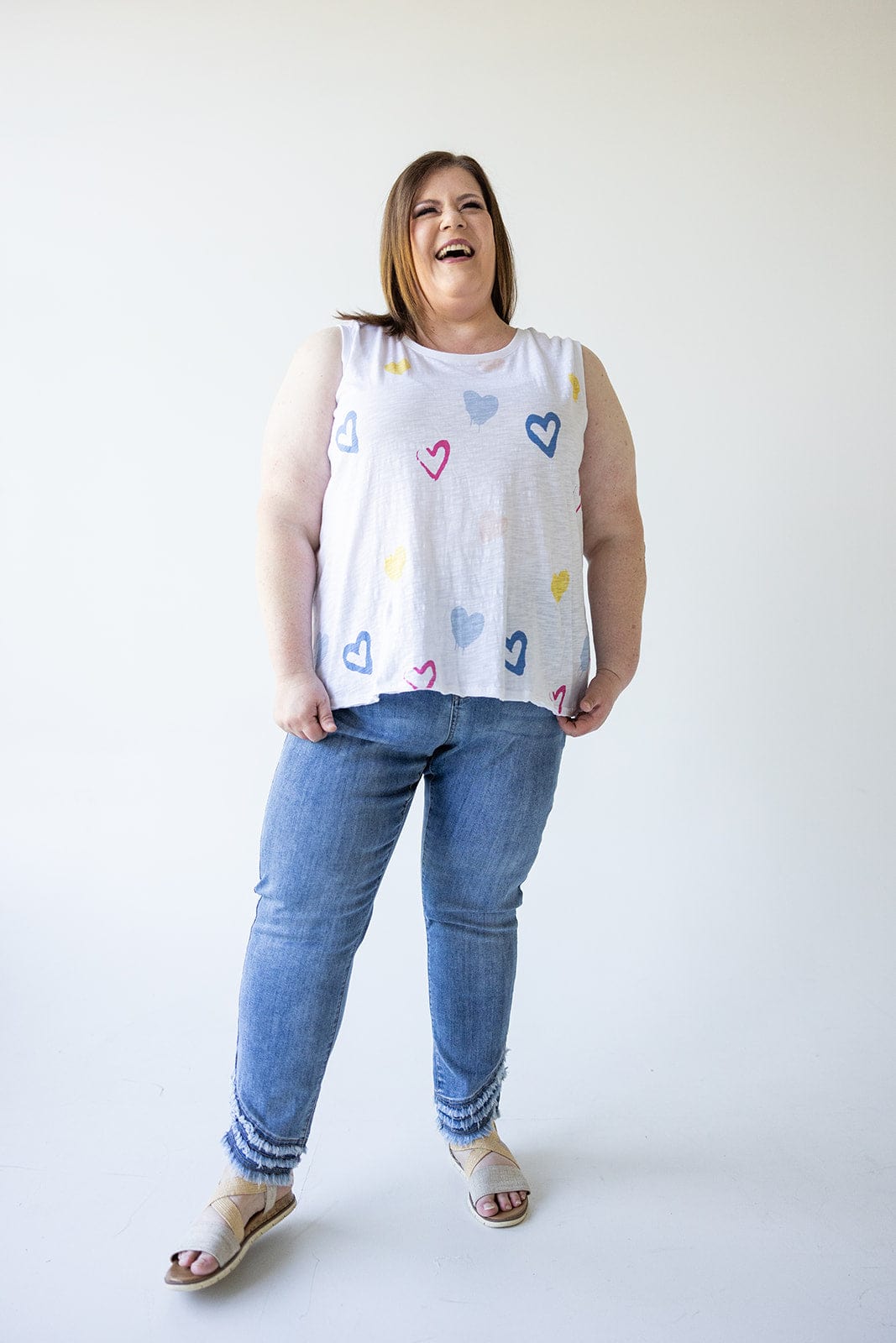 A person dressed in a sleeveless shirt adorned with colorful heart designs is standing and smiling against a plain background, wearing Love Marlow’s High-Rise Relaxed Skinny Jeans with Frayed Hem in Medium Wash.