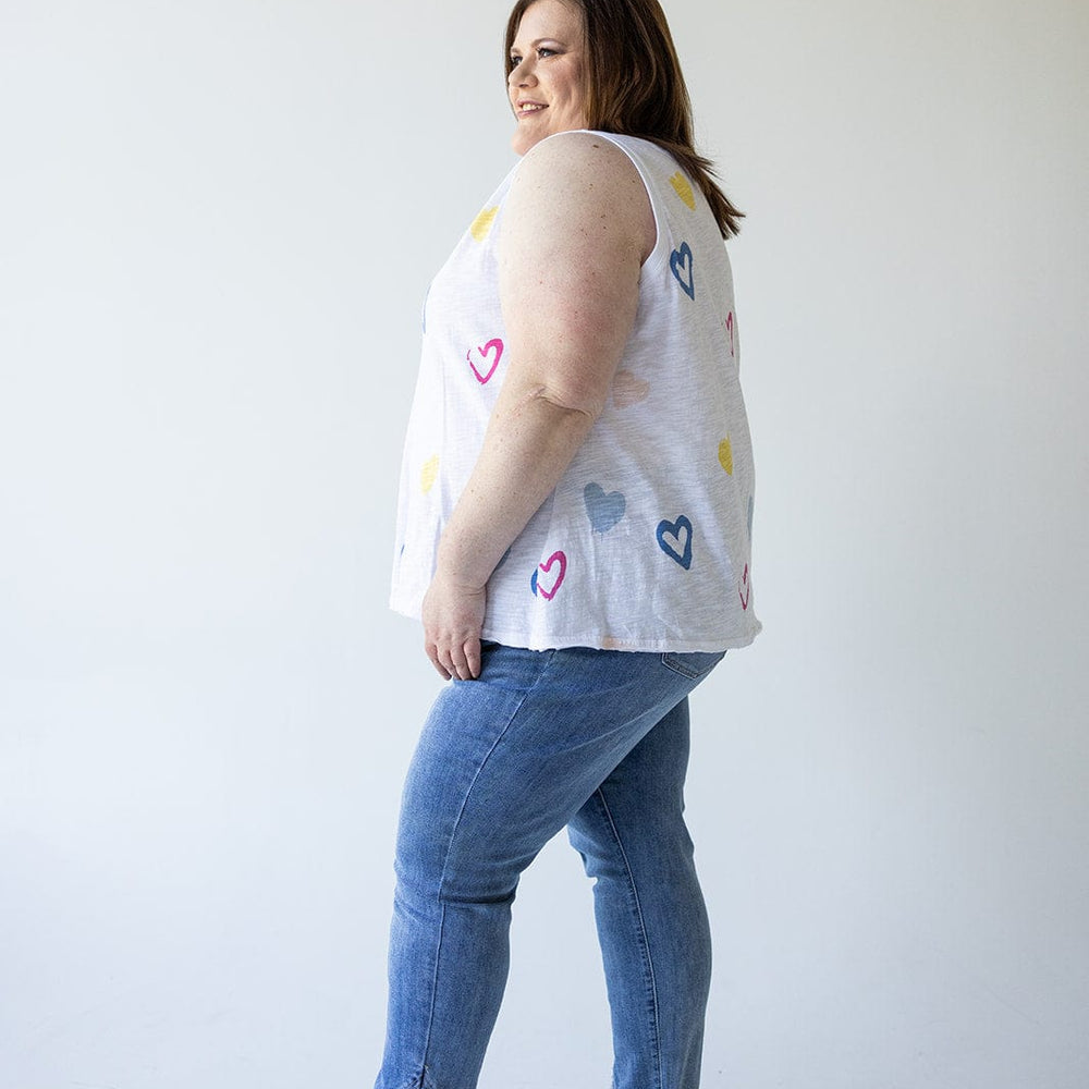 A person stands in profile facing left, wearing a sleeveless top adorned with heart patterns and the HIGH-RISE RELAXED SKINNY JEAN WITH FRAYED HEM IN MEDIUM WASH by Love Marlow, paired with sandals against a plain background.