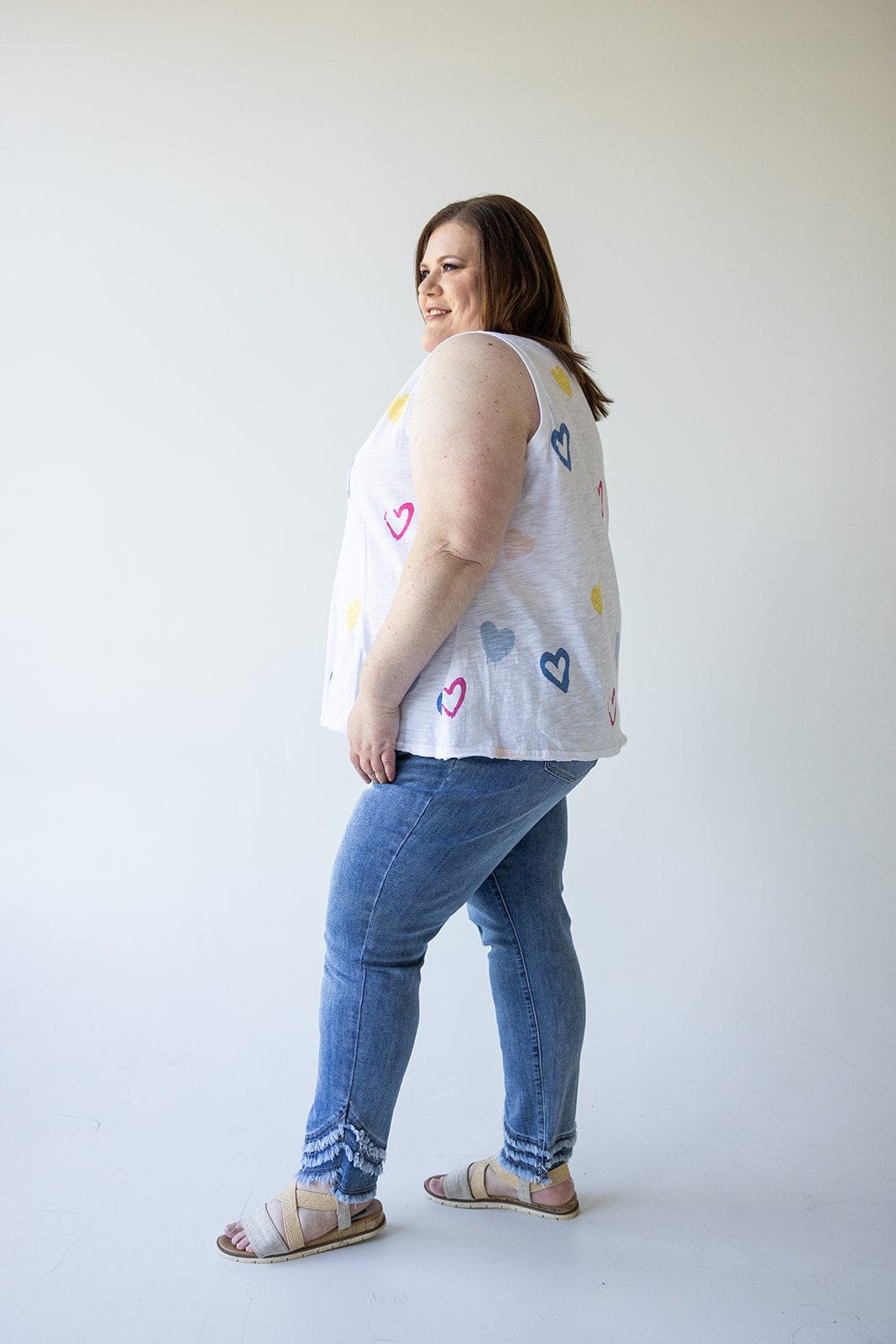 A person stands in profile facing left, wearing a sleeveless top adorned with heart patterns and the HIGH-RISE RELAXED SKINNY JEAN WITH FRAYED HEM IN MEDIUM WASH by Love Marlow, paired with sandals against a plain background.