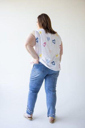 In a light-filled room, a person stands with their back to the camera, wearing a white top featuring colorful heart patterns and Love Marlow's High-Rise Relaxed Skinny Jeans with Frayed Hem in Medium Wash.