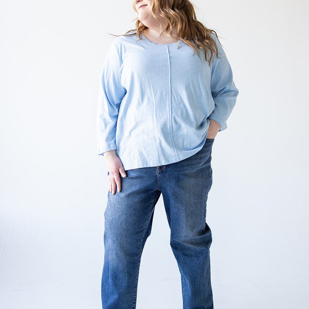 A person in a light blue shirt and relaxed skinny jeans stands with one hand in their pocket, looking to the side. The background is plain white.