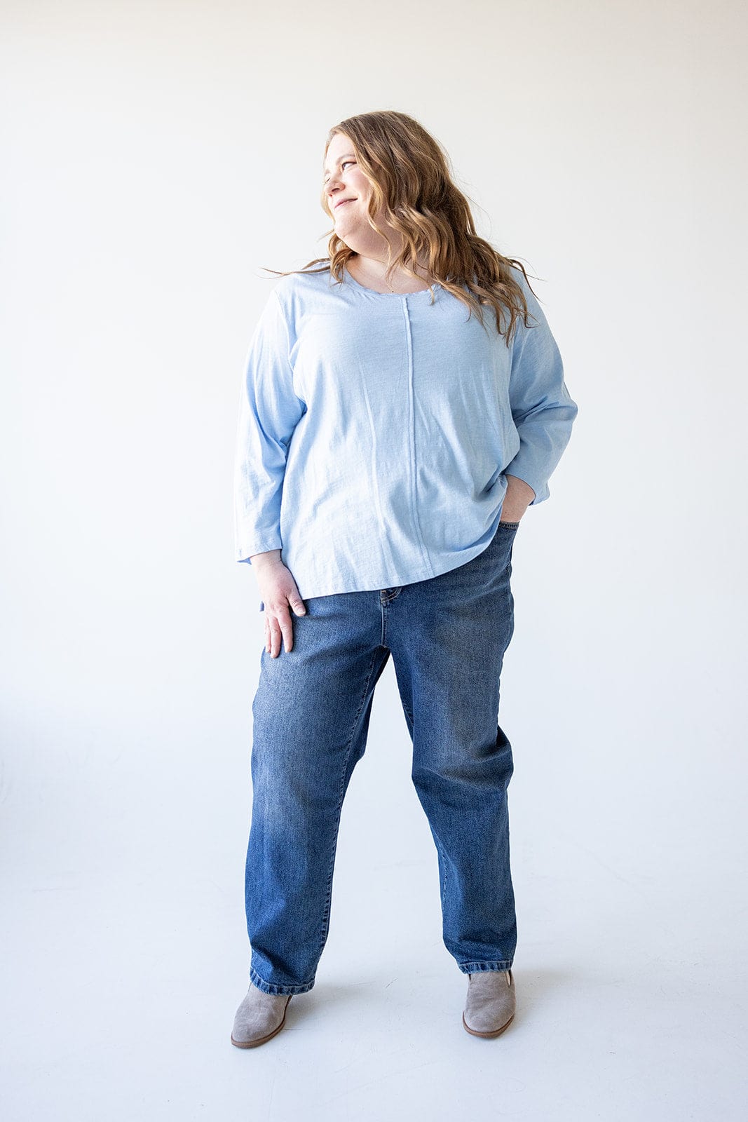 A person in a light blue shirt and relaxed skinny jeans stands with one hand in their pocket, looking to the side. The background is plain white.