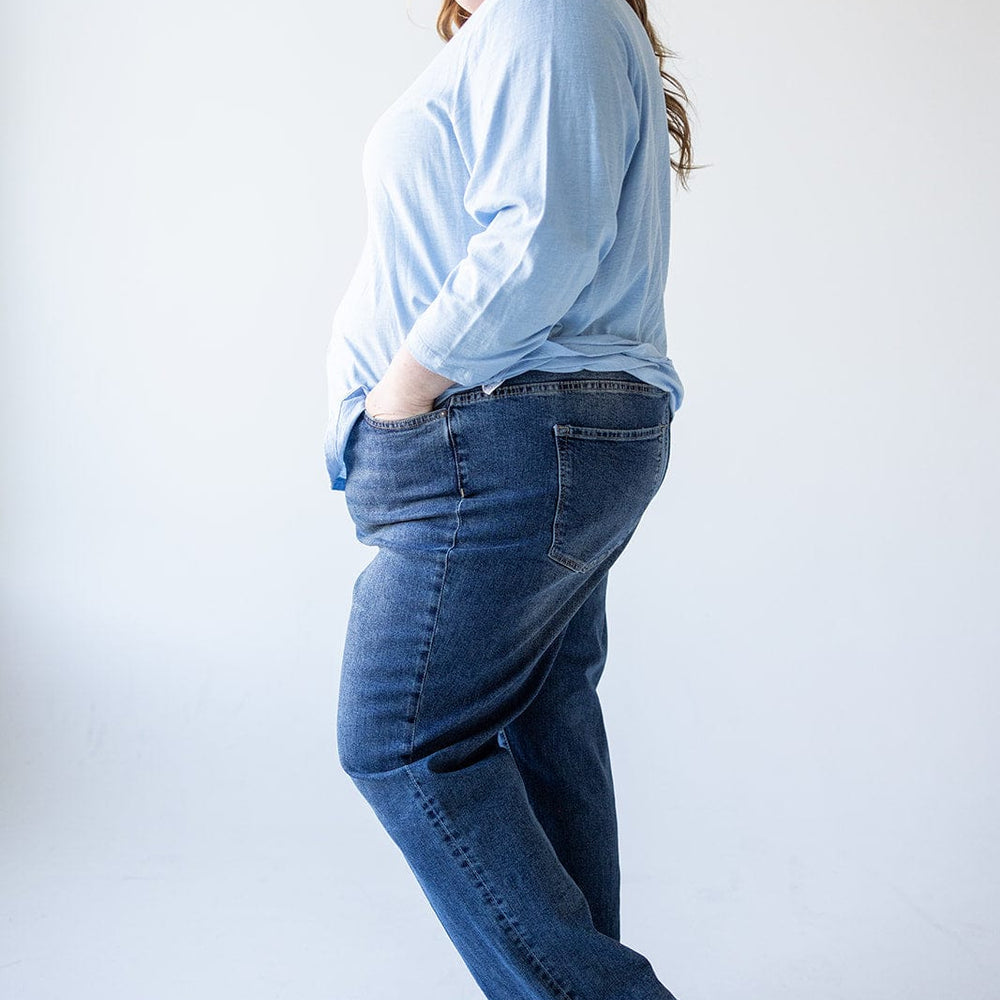 A woman in a light blue shirt and high-rise, relaxed skinny jeans stands sideways with hands in pockets, smiling against a plain background.