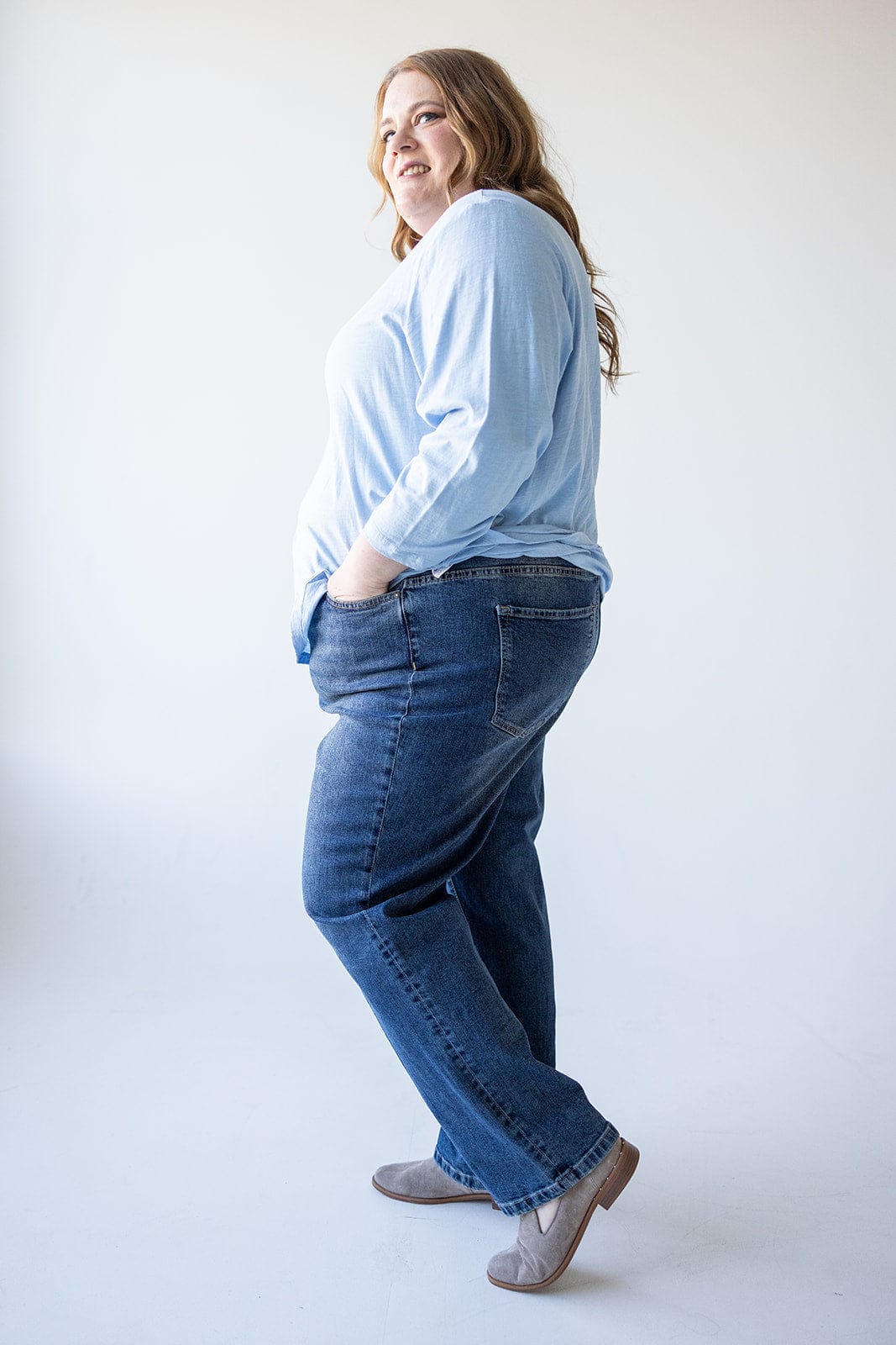 A woman in a light blue shirt and high-rise, relaxed skinny jeans stands sideways with hands in pockets, smiling against a plain background.