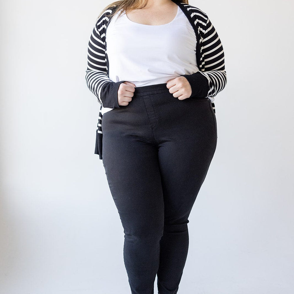 A woman stands against a white background wearing a white top, Love Marlow HIGH-RISE SKINNY JEGGING IN BLACK, a black and white striped cardigan, and blue sandals.