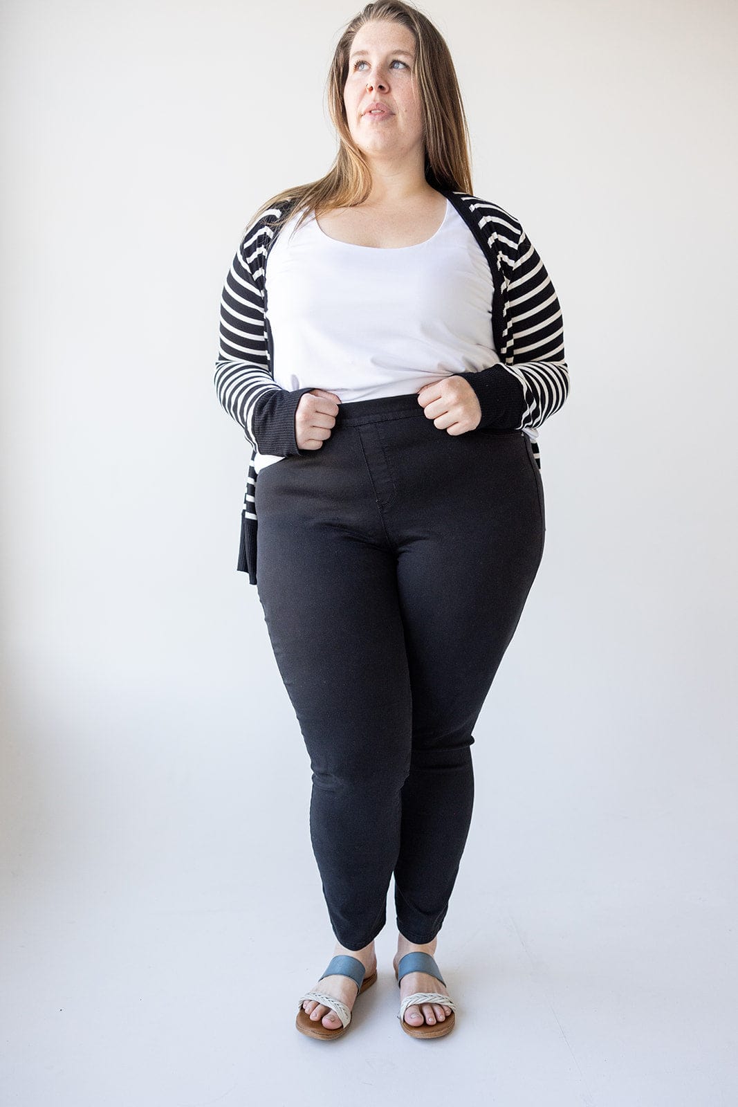 A woman stands against a white background wearing a white top, Love Marlow HIGH-RISE SKINNY JEGGING IN BLACK, a black and white striped cardigan, and blue sandals.