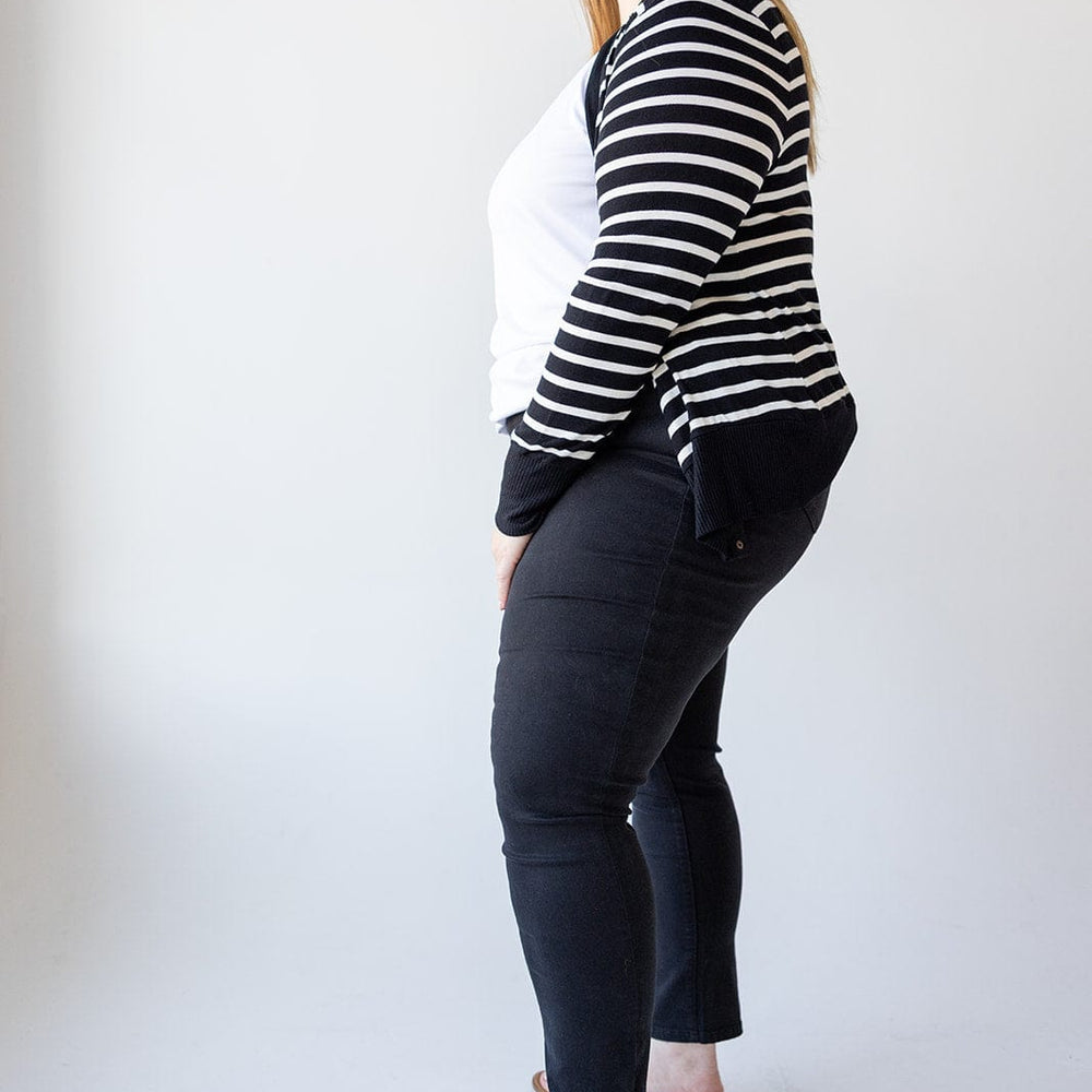 A woman with long hair stands in profile wearing a black and white striped cardigan, white shirt, Love Marlow HIGH-RISE SKINNY JEGGING IN BLACK, and sandals against a plain background.