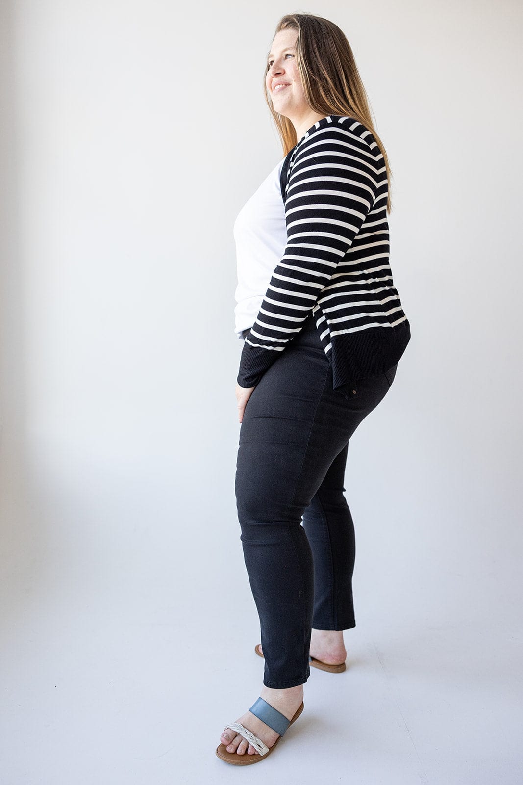 A woman with long hair stands in profile wearing a black and white striped cardigan, white shirt, Love Marlow HIGH-RISE SKINNY JEGGING IN BLACK, and sandals against a plain background.