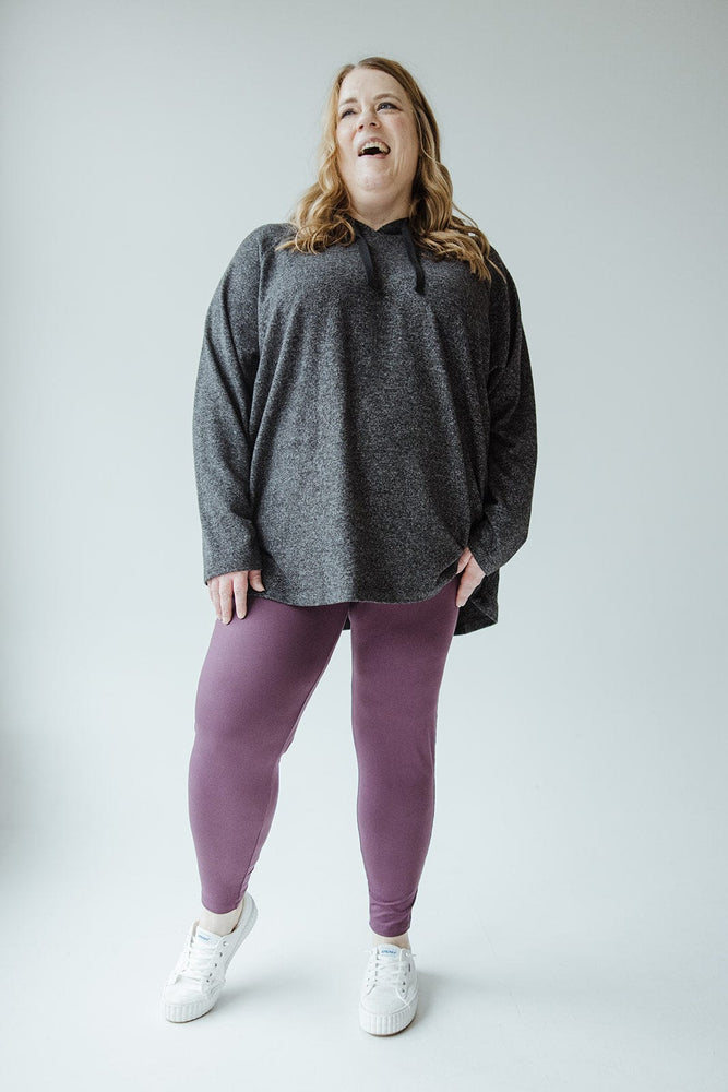 A person with long hair wearing a gray hoodie, Zenana High-Waisted Super Soft Leggings in Antique Violet, and white sneakers smiles while standing against a plain background.