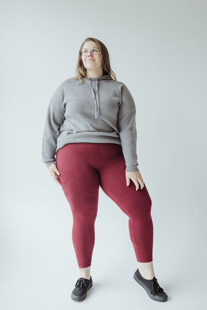 A person stands against a plain background, wearing a grey sweatshirt, HIGH-WAISTED SUPER SOFT LEGGINGS IN DEEP BURGUNDY by Zenana, and black shoes.