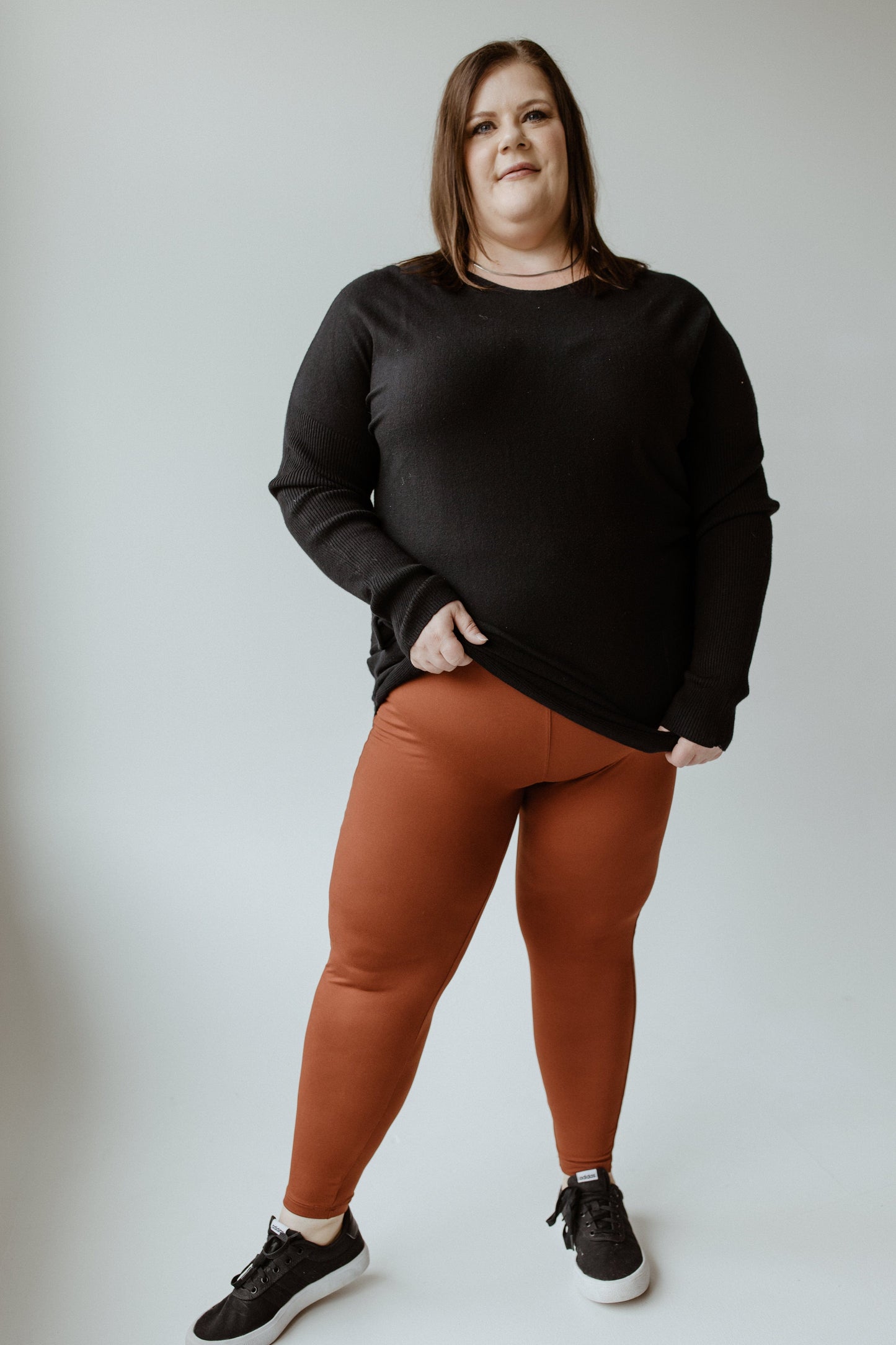 A woman stands against a plain background, wearing a black sweater and ultra-soft, high-waisted leggings paired with black sneakers.