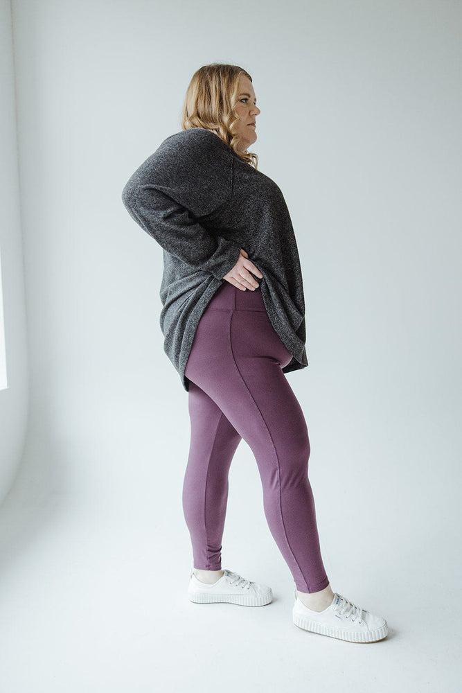 A person with long hair stands facing away, wearing a dark gray hoodie, Zenana's High-Waisted Super Soft Leggings in Antique Violet, and white sneakers, against a plain, light-colored background.