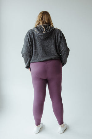 A person with long hair stands facing away, wearing a dark gray hoodie, Zenana's High-Waisted Super Soft Leggings in Antique Violet, and white sneakers, against a plain, light-colored background.