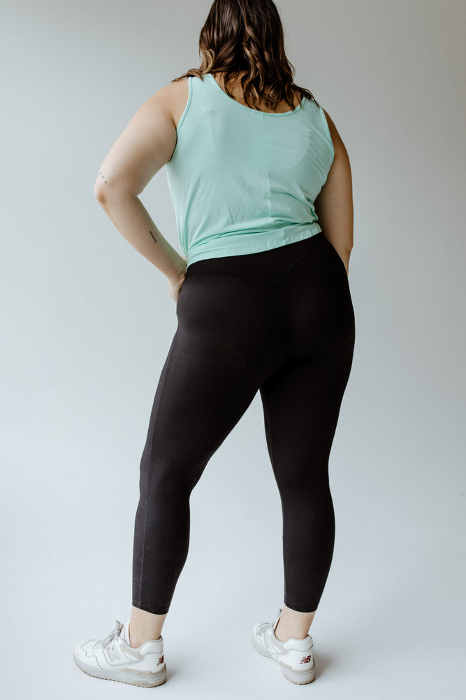 A person in a mint green tank top, Zenana's HIGH-WAISTED SUPER SOFT LEGGINGS IN BLACK, and white sneakers stands with hands on hips against a plain background.