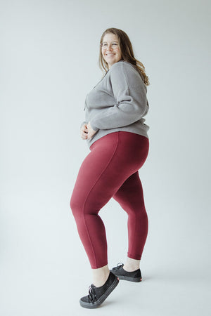 A woman stands smiling in a light gray sweater, Zenana HIGH-WAISTED SUPER SOFT LEGGINGS IN DEEP BURGUNDY, and black shoes against a plain white background.