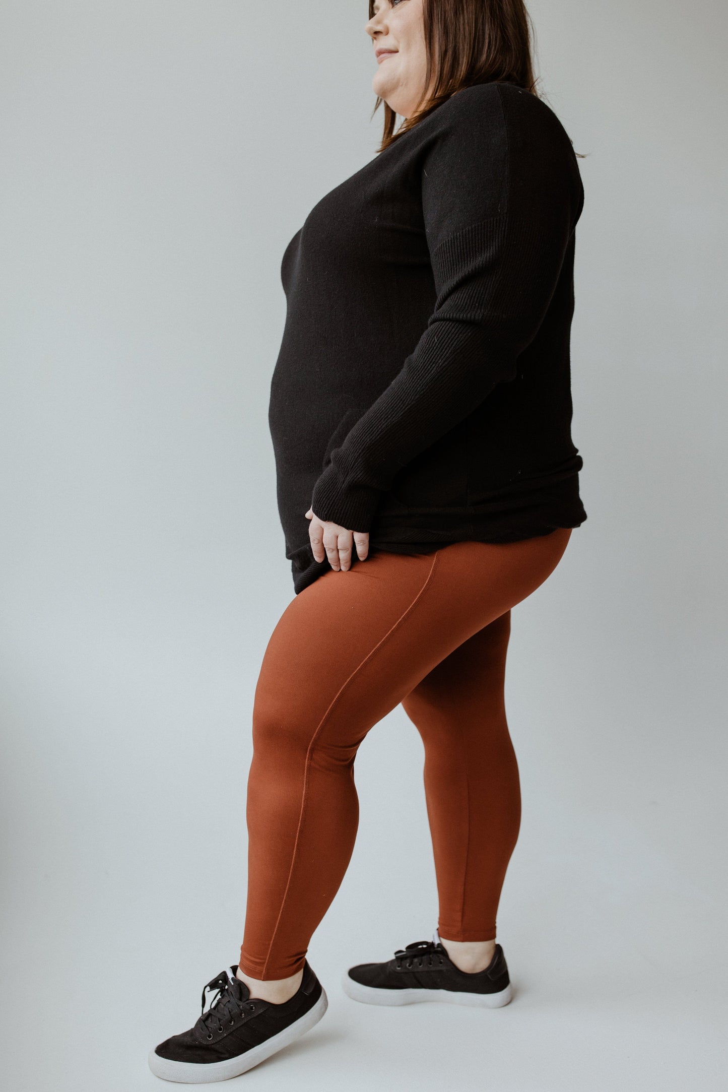 A person wearing a black sweater, rust-colored ultra-soft leggings, and black sneakers stands against a plain background.