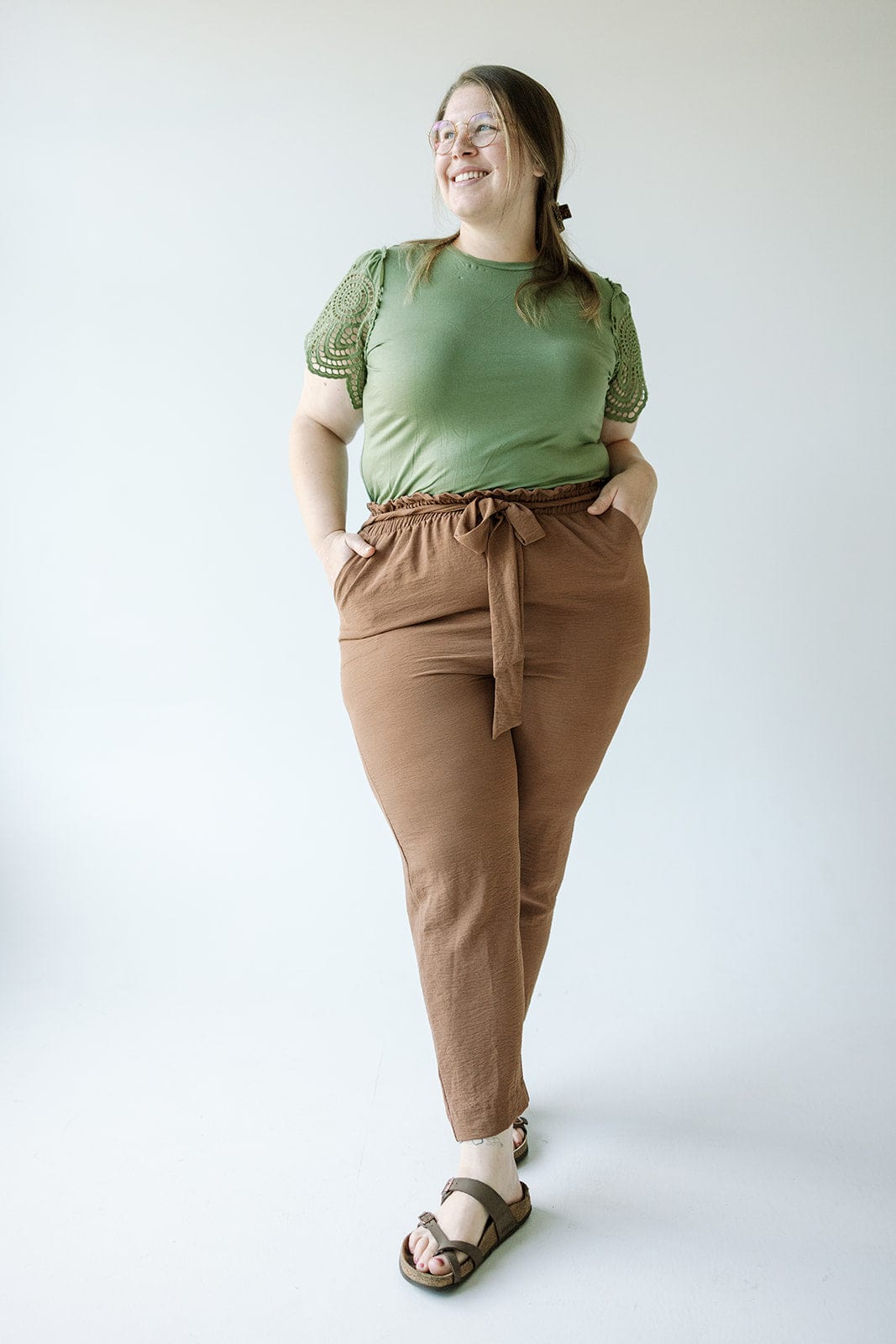 A woman wearing a green top and the HIGH-WAIST PAPERBAG PANT IN SALTED CARMEL by Love Marlow, along with sandals, stands against a plain background, smiling with her hands in her pockets.