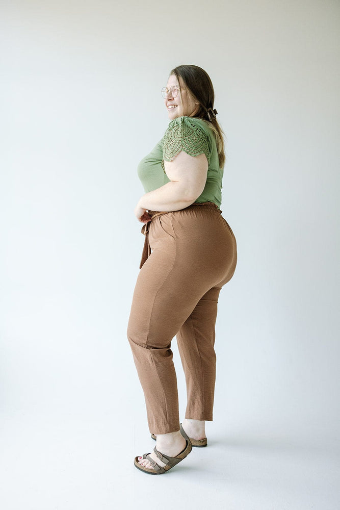 A woman with light brown hair wearing a green short-sleeve top, Love Marlow's High-Waist Paperbag Pant in Salted Caramel, and brown shoes is standing with her back to the camera.