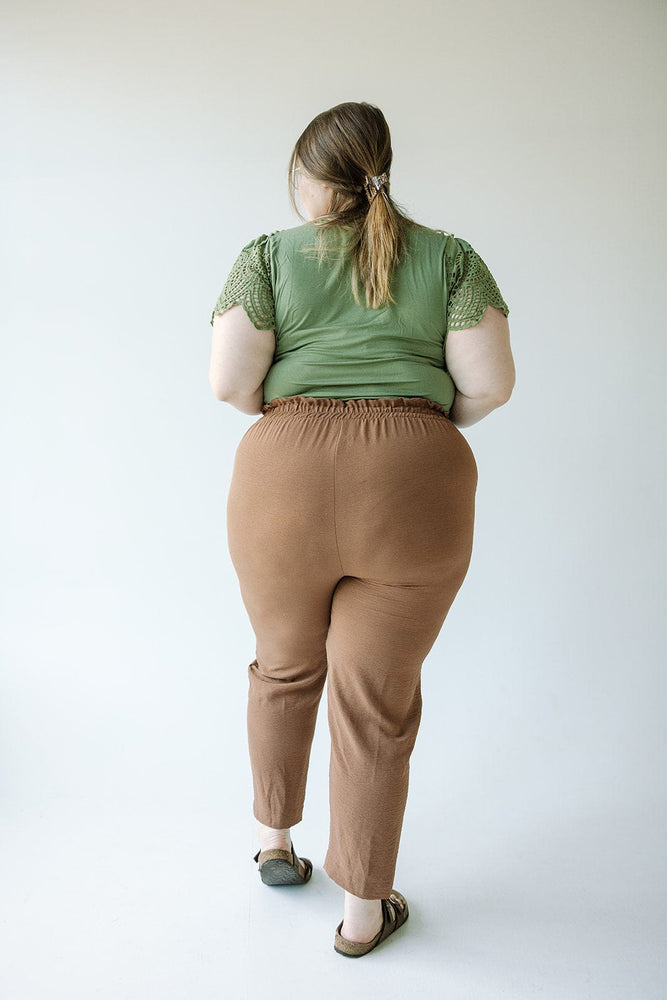 A woman with light brown hair wearing a green short-sleeve top, Love Marlow's High-Waist Paperbag Pant in Salted Caramel, and brown shoes is standing with her back to the camera.