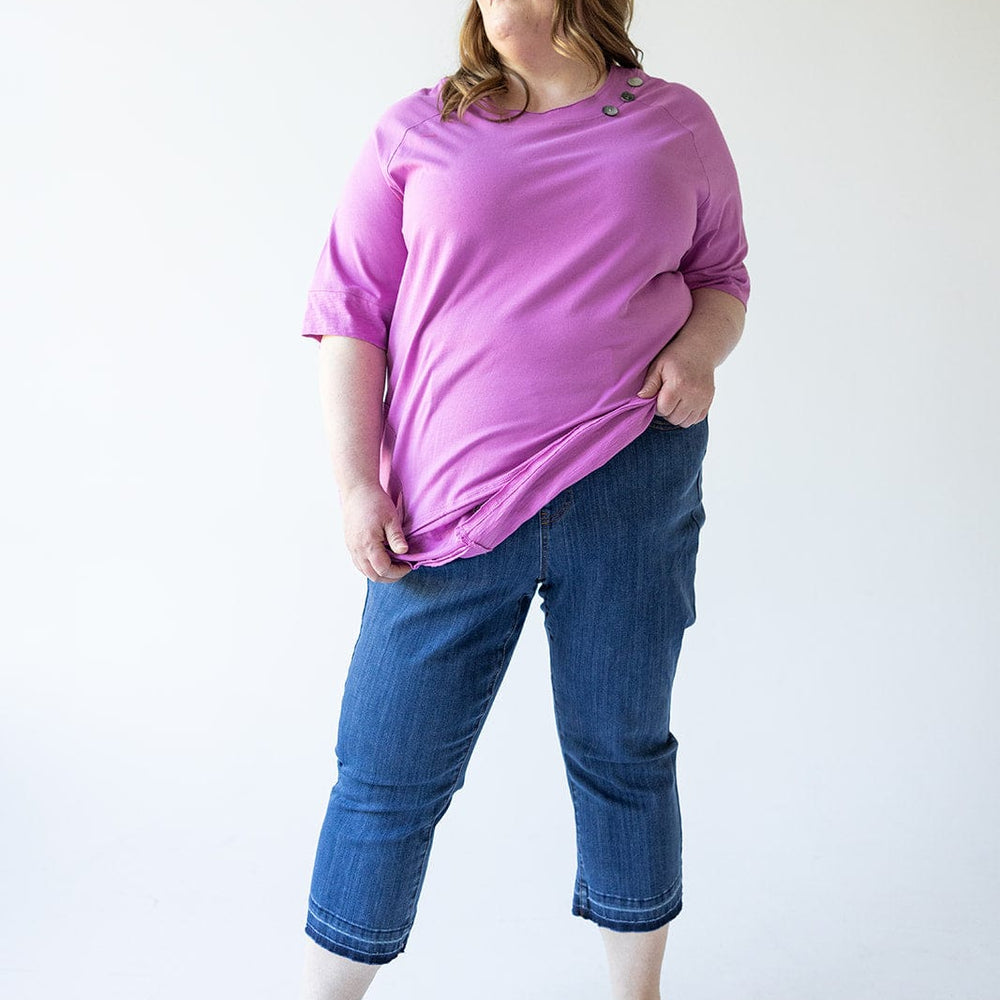 A woman stands against a plain background wearing a purple top, Judy Blue High Waist Pull On Capri with Pockets jeans, and brown sandals, with her left hand in her pocket and looking off to her left.