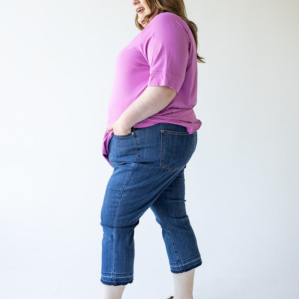 A person stands in a casual pose, wearing a pink shirt, Judy Blue High Waist Pull On Capri with Pockets, and sandals, against a plain background.