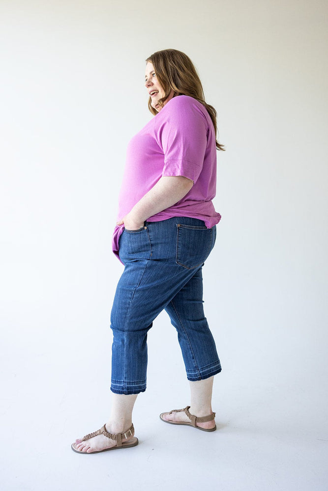 A person stands in a casual pose, wearing a pink shirt, Judy Blue High Waist Pull On Capri with Pockets, and sandals, against a plain background.