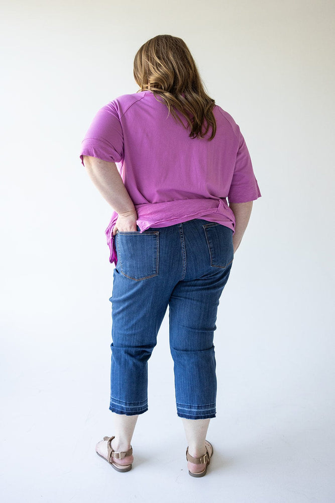 A person with shoulder-length hair wearing a pink top, Judy Blue High Waist Pull On Capri with Pockets jeans, and sandals stands facing away, with hands on their hips.