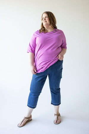 A woman stands against a plain background wearing a purple top, Judy Blue High Waist Pull On Capri with Pockets jeans, and brown sandals, with her left hand in her pocket and looking off to her left.