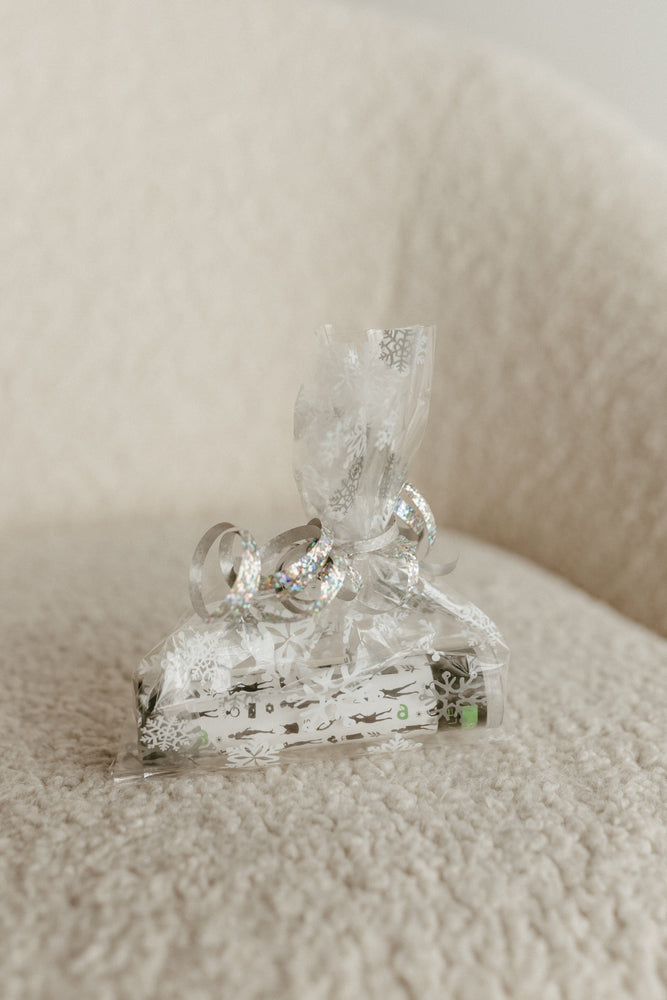 A small gift wrapped in clear snowflake-patterned paper, secured with fashion tape for a strong hold, and tied with a shiny ribbon, sits elegantly on a textured beige fabric surface.